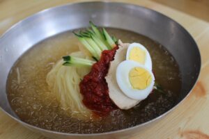 冷たいスープに麺、キュウリ、ゆで卵と赤い薬味を盛った水ミルミョン